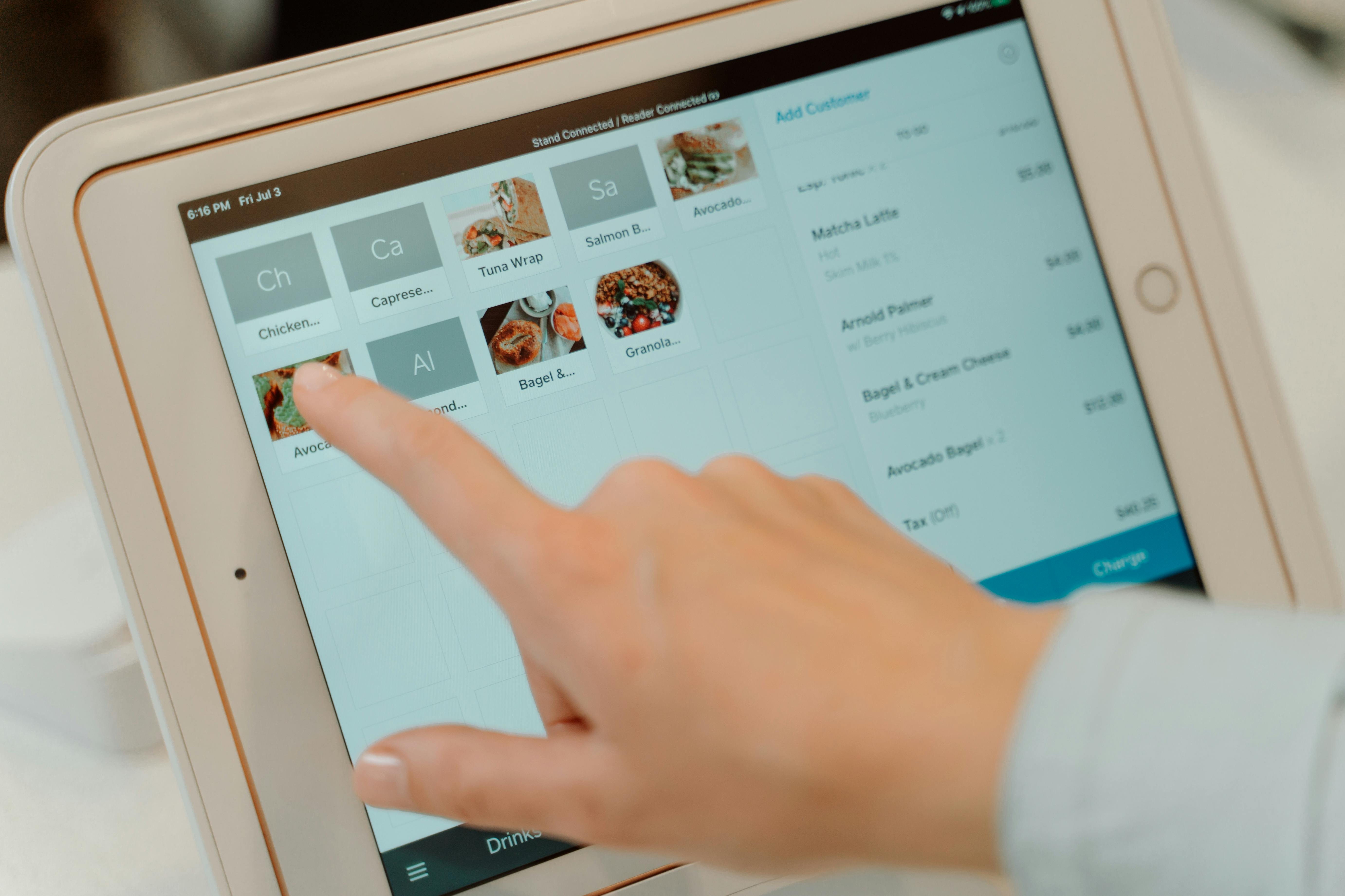 A finger tapping a menu item on a tablet POS screen showing a café order